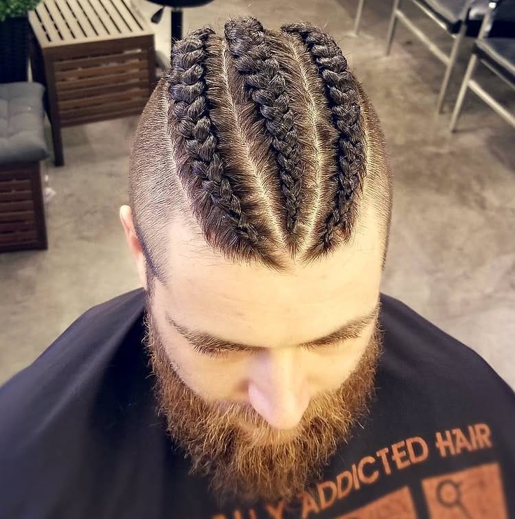 A bearded white man with thick brown hair styled into three large, voluminous Dutch braids and a sharp disconnected undercut.