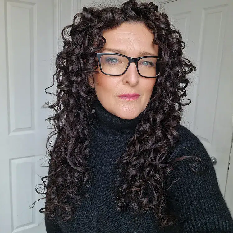 Older woman with long dark curly hair, voluminous spiral curls, and black rimmed glasses.