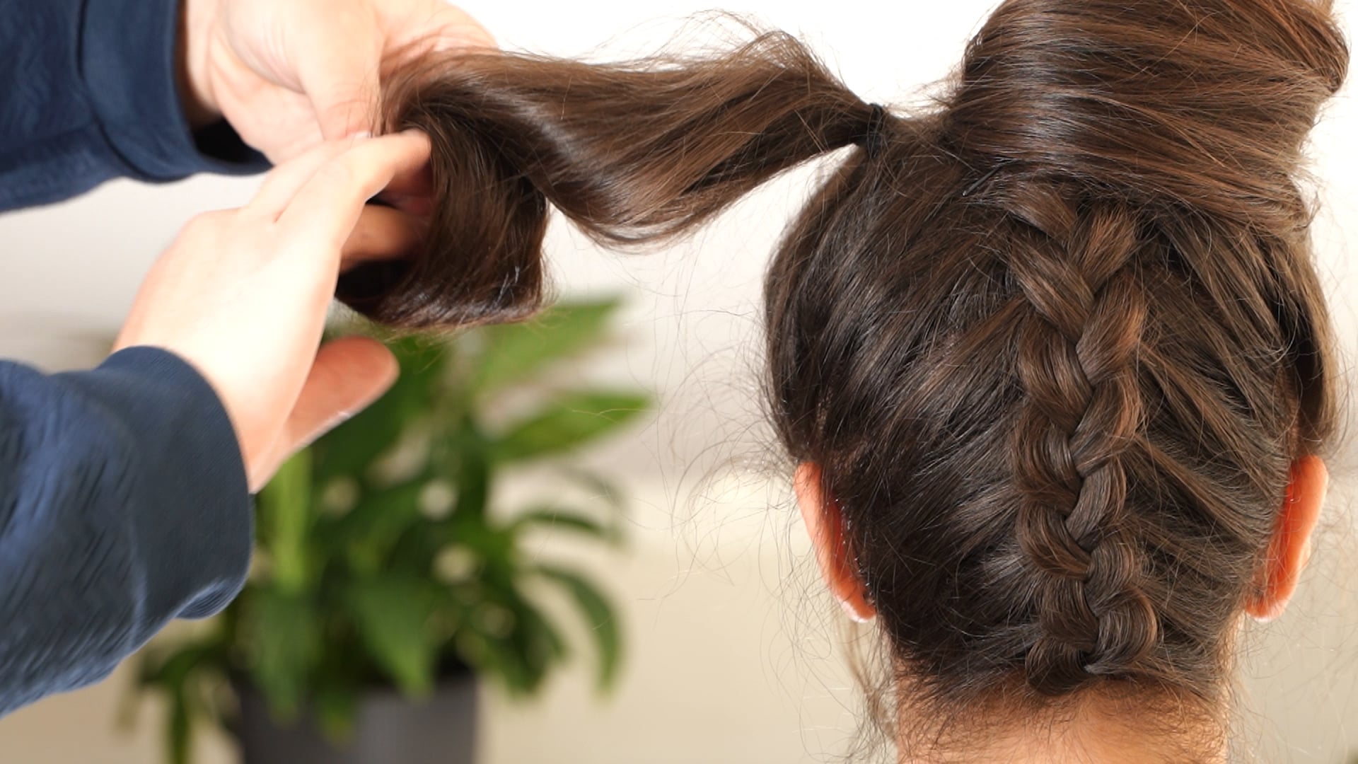 Back view of a hairstyle with an upside-down center braid as a stylist rolls one side section inward to form one loop of the bow.