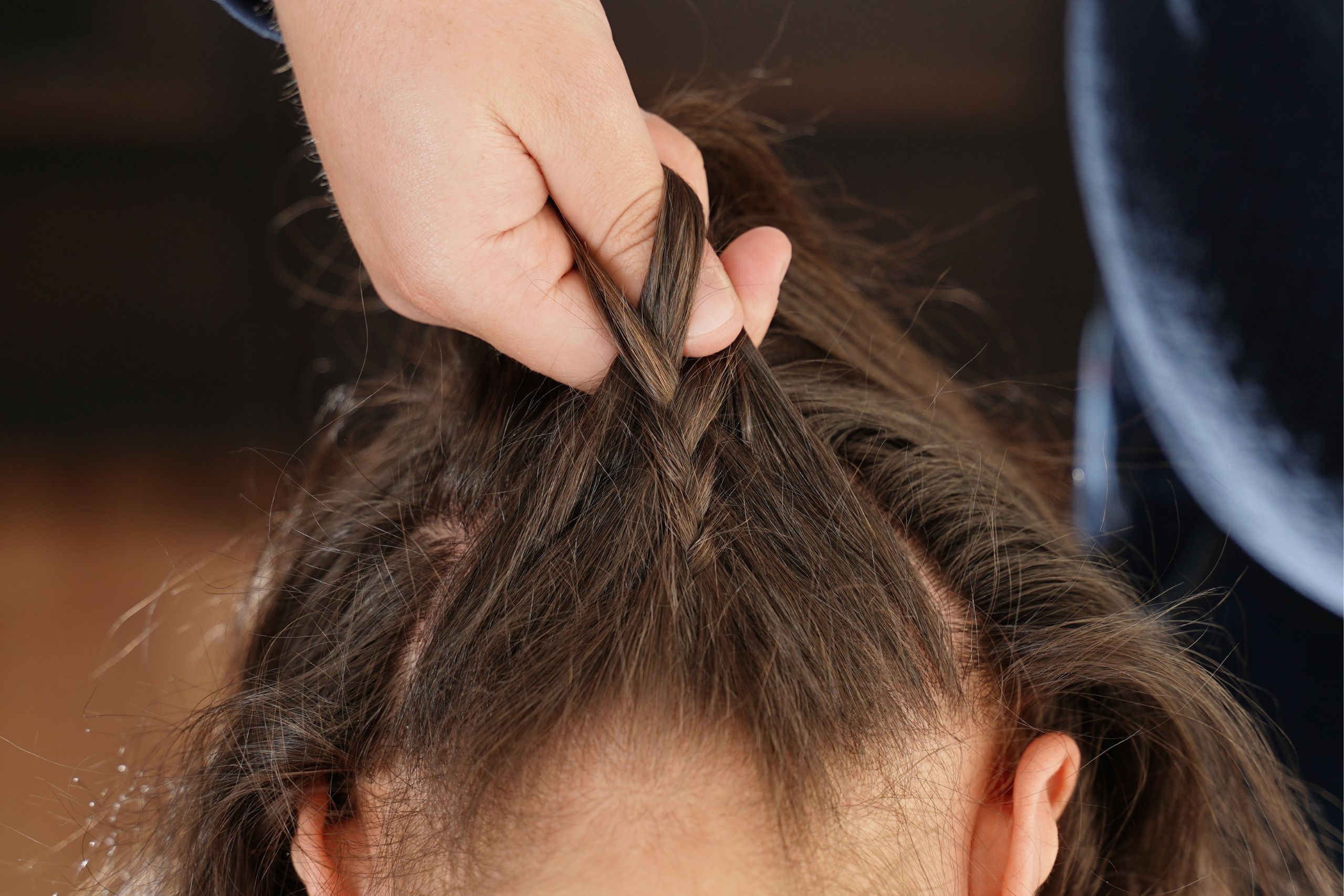 Hand starting an upside-down Dutch braid at the nape while the person&rsquo;s head is tilted forward.