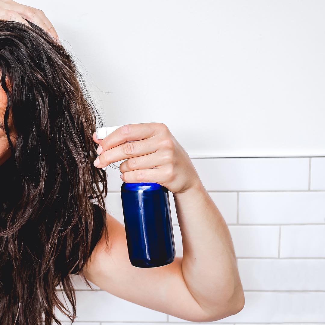 Woman applying a homemade aloe and glycerin leave in spray from a blue glass spray bottle to damp hair