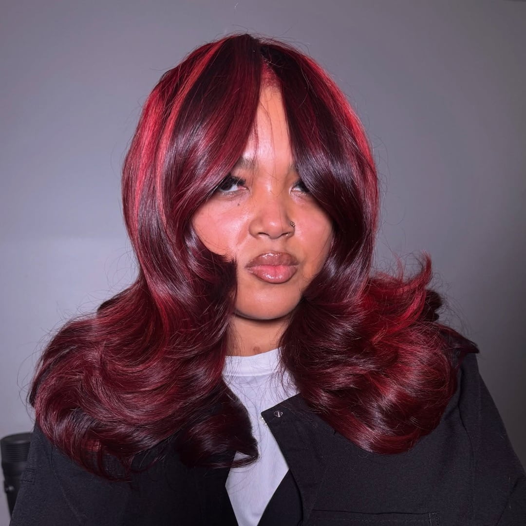 Voluminous burgundy and red highlighted blowout hair featuring center-parted curtain bangs