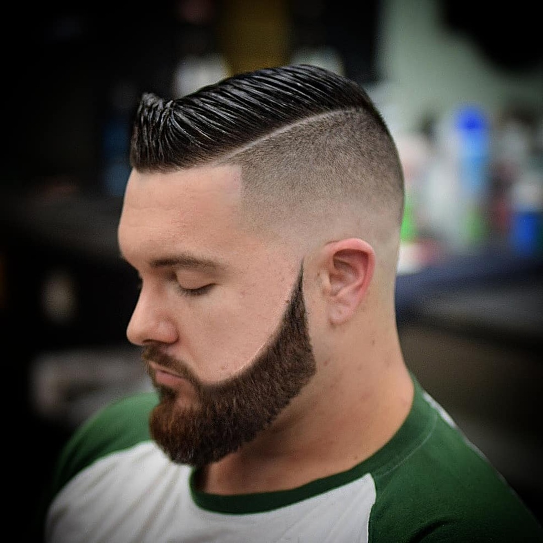 Man with a high skin fade comb over featuring a raised front quiff, sharp hard part, and full shaped beard